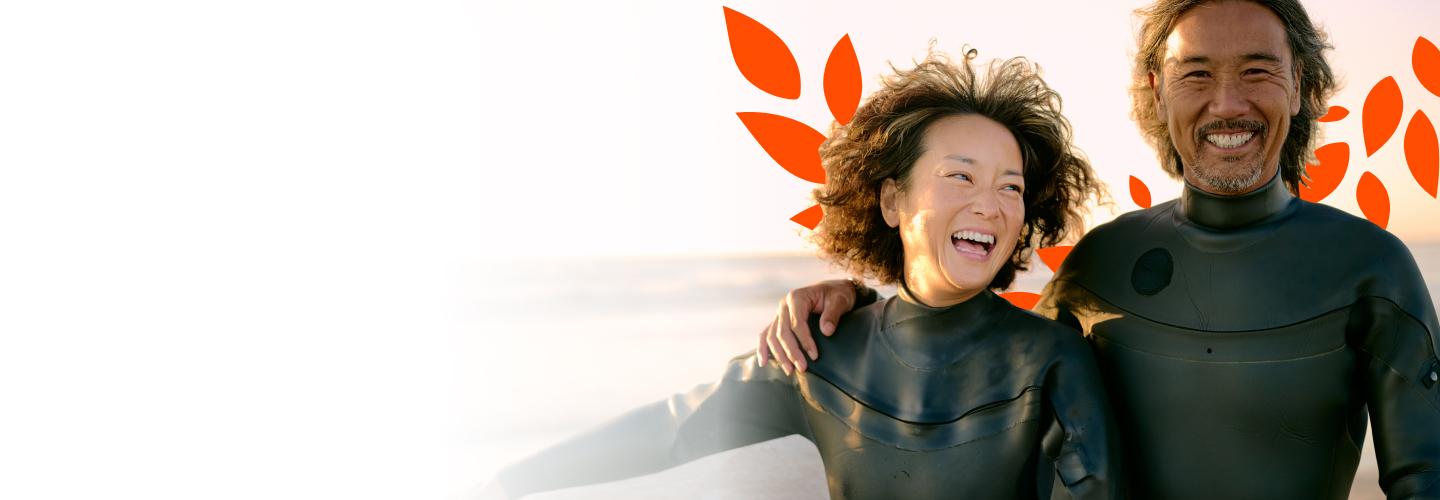 A man and a woman in wetsuits smile at the beach